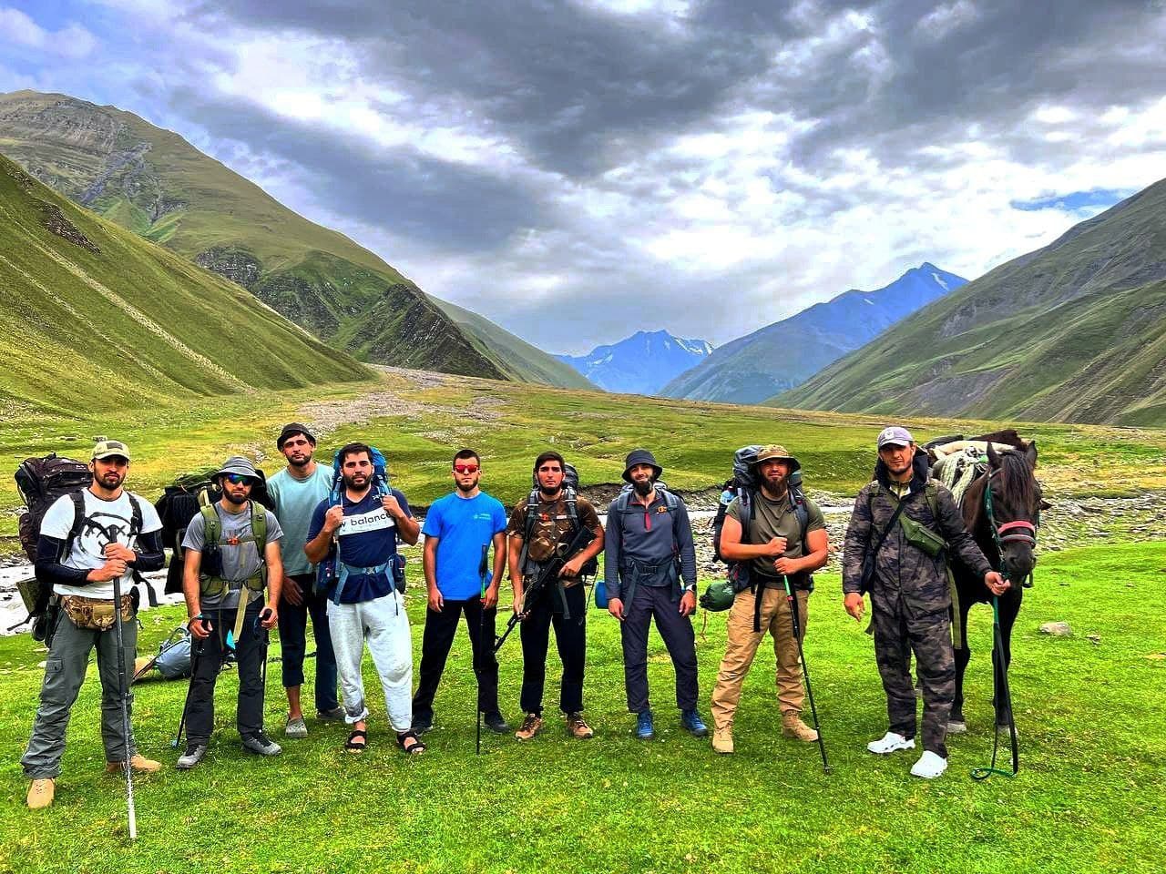 A DIY Trails group standing with packs and trekking poles on a high meadow, ridgelines and snow peaks behind them